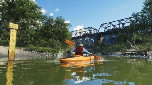 A kayaker paddles toward a rail bridge as a train crosses, a flood gauge beside him.