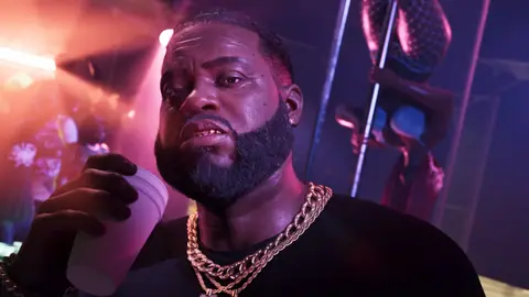 In the club with a foam cup, gold chains and gold teeth, a dancer on the pole behind him.