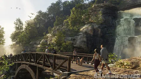 A wooden footbridge beside a waterfall in a wooded park, with people and a dog crossing.