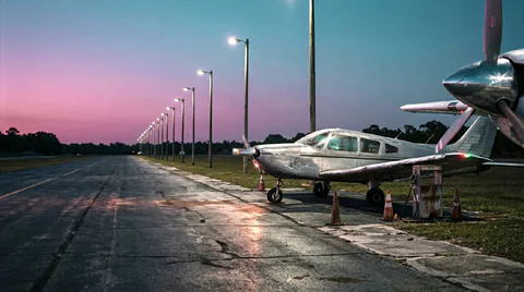 Small aircraft are parked beside a runway lined with lights beneath a pink and blue evening sky.