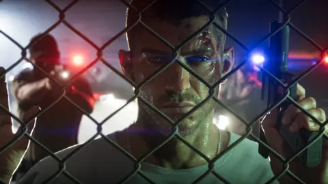 Behind a chain-link fence at night with a cut on his brow, police lights behind him.