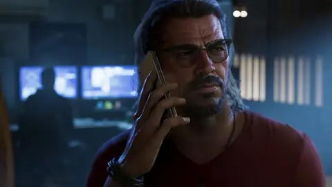 On the phone in glasses while someone works at two computer monitors behind him.