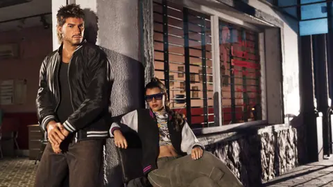 Jason in a black leather jacket and Lucia in sunglasses and a varsity jacket outside a shop.
