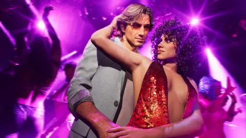 Jason in tinted glasses dances with Lucia in the red sequin dress under purple club lights.